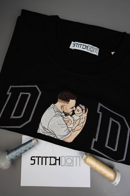 Tricou negru din bumbac organic, împăturit, cu o poză brodată personalizată cu un tată care își ține copilul în brațe, încadrat de două litere mari „D” conturate cu gri, alături de conuri de ață și o etichetă albă cu brandul „STITCHDOM”, pe un fundal gri.