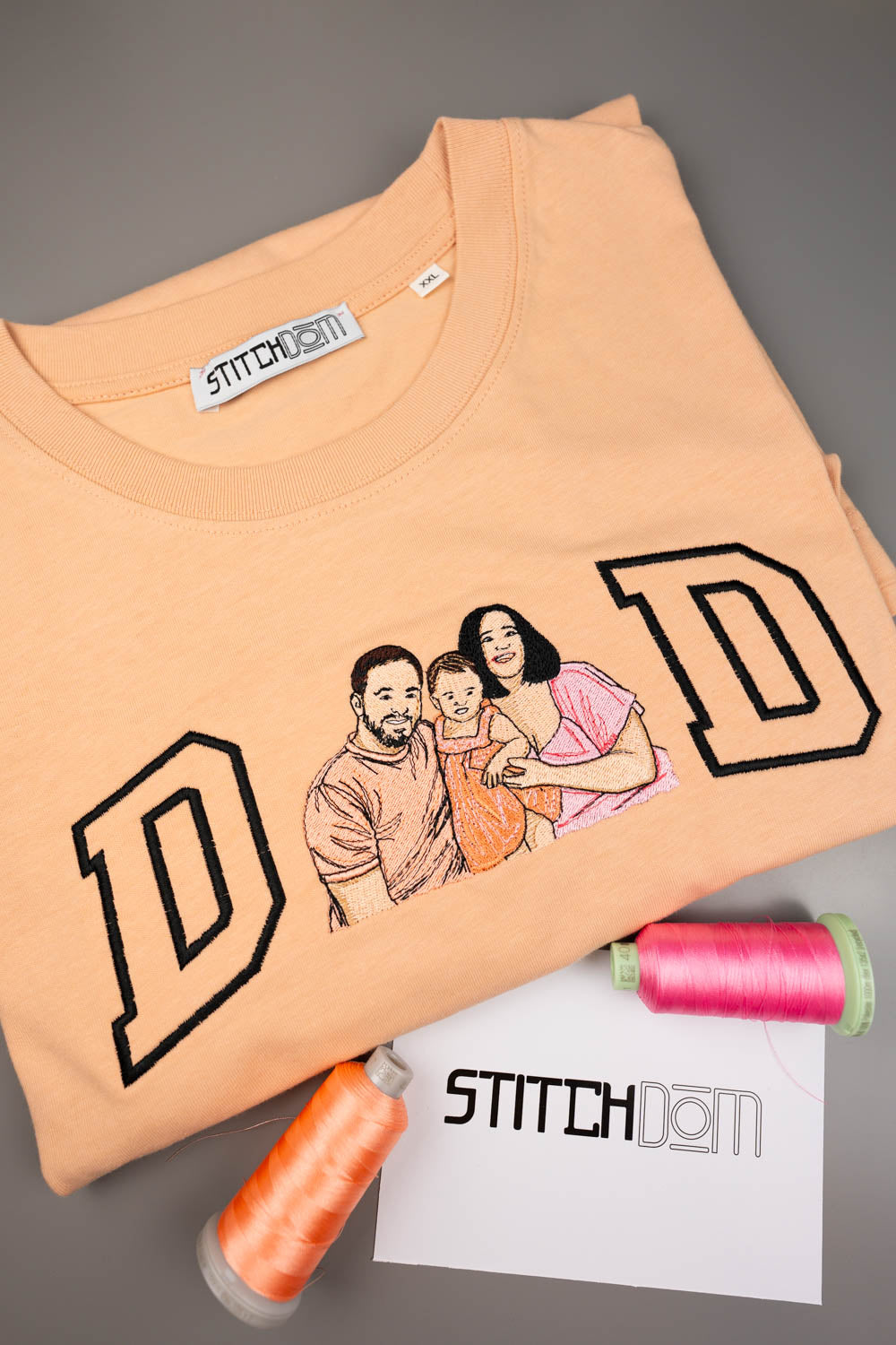 Tricou piersică din bumbac organic, cu o poză brodată personalizată cu o familie și textul „DAD” realizat din litere mari conturate cu negru, alături de conuri de ață și un card „Stitchdom”, pe un fundal gri.