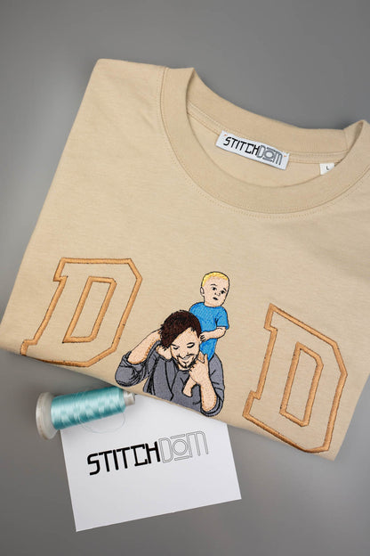 Tricou bej din bumbac organic, împăturit, cu o poză brodată personalizată cu un bărbat și un copil, încadrată de două litere mari „D” conturate, alături de un con de ață bleu și un card „Stitchdom”, pe un fundal gri.
