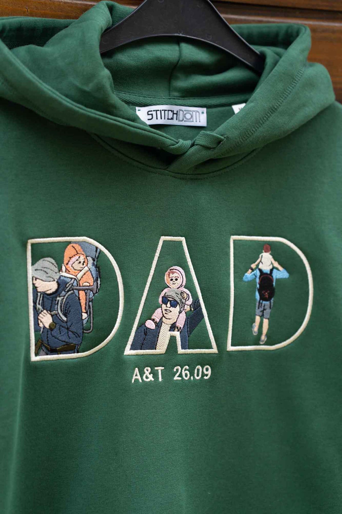 Hanorac verde cu glugă, pe umeraș, cu designul „DAD” brodat, fiecare literă conținând elemente brodate.