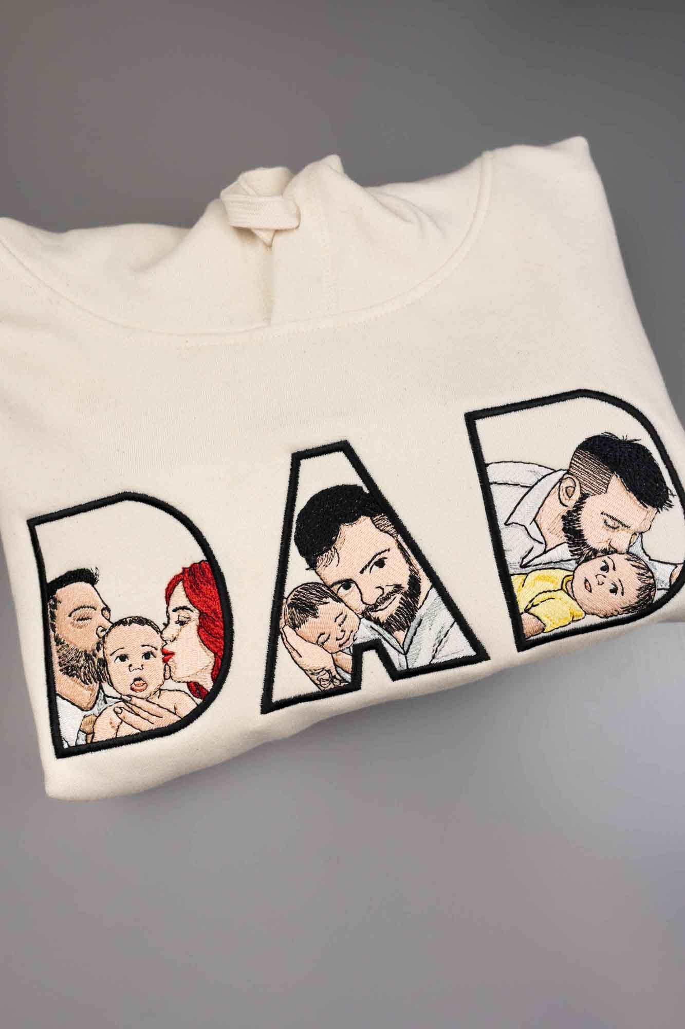 Hanorac alb cu glugă, cu textul „DAD” brodat, fiecare literă conținând imagini brodate reprezentând un tată și un copil, pe un fundal gri.