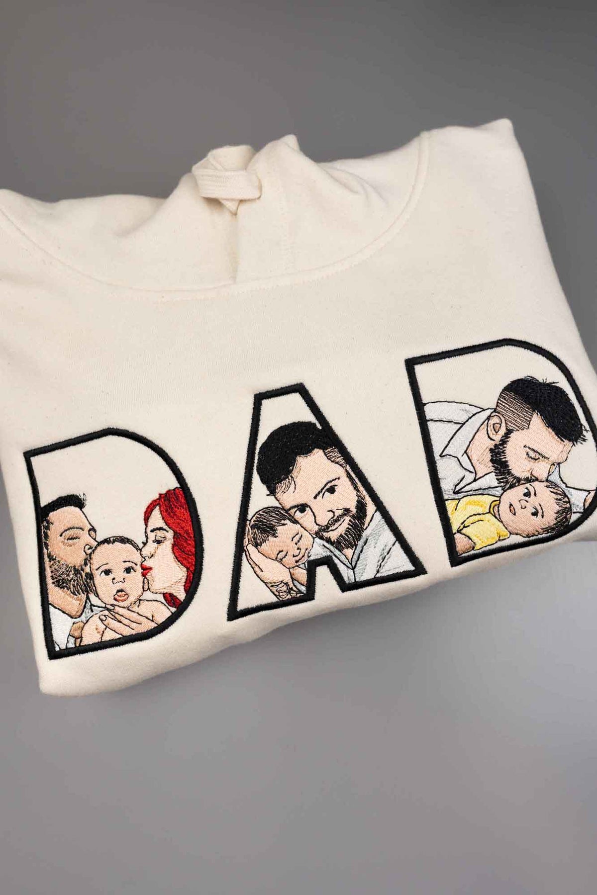 Hanorac alb cu glugă, cu textul „DAD” brodat, fiecare literă conținând imagini brodate reprezentând un tată și un copil, pe un fundal gri.