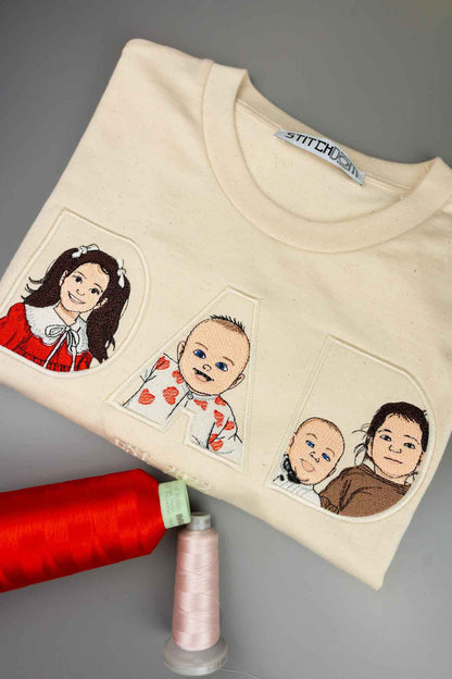 Tricou crem împăturit cu textul „DAD” brodat, fiecare literă conținând câte un portret brodat al copiilor, așezat pe un fundal gri, alături de două conuri de ață.