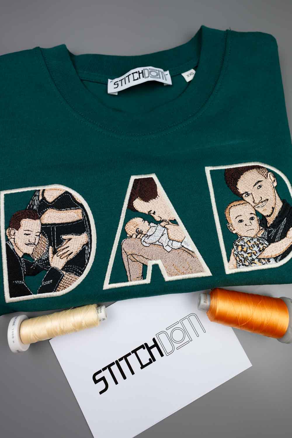 Tricou verde petrol închis din bumbac organic, împăturit, cu textul „DAD” realizat din litere brodate mari, cu poze de familie în interiorul literelor, eticheta „Stitchdom” vizibilă la guler și două conuri de ață cu capete alb și portocaliu, alături de un card „Stitchdom”, pe o suprafață gri.
