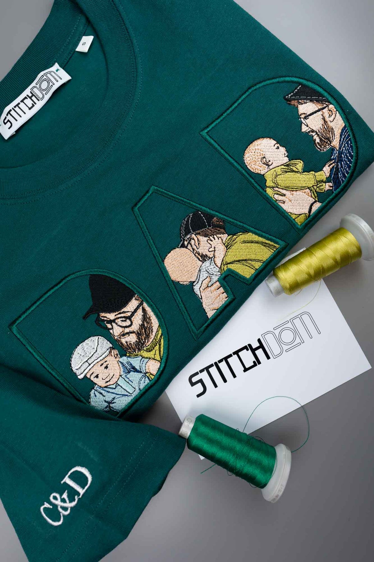 Tricou verde petrol închis din bumbac organic, cu poze brodate în stil desen cu tați și copii pe piept, alături de conuri de ață și un card de brand, pe un fundal gri.