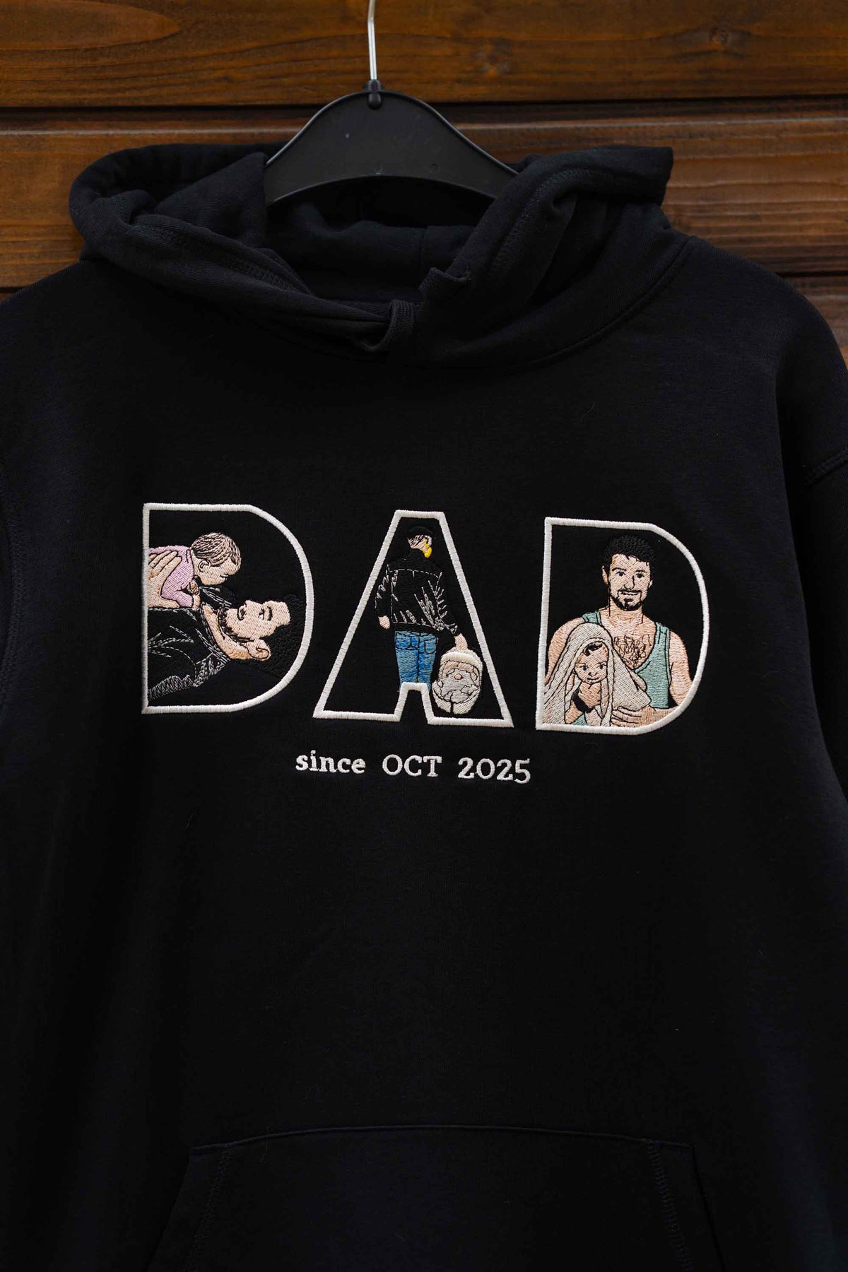 Hanorac negru cu glugă, cu textul „DAD since OCT 2025” brodat, fiecare literă conținând elemente brodate, pe fundal din lemn.