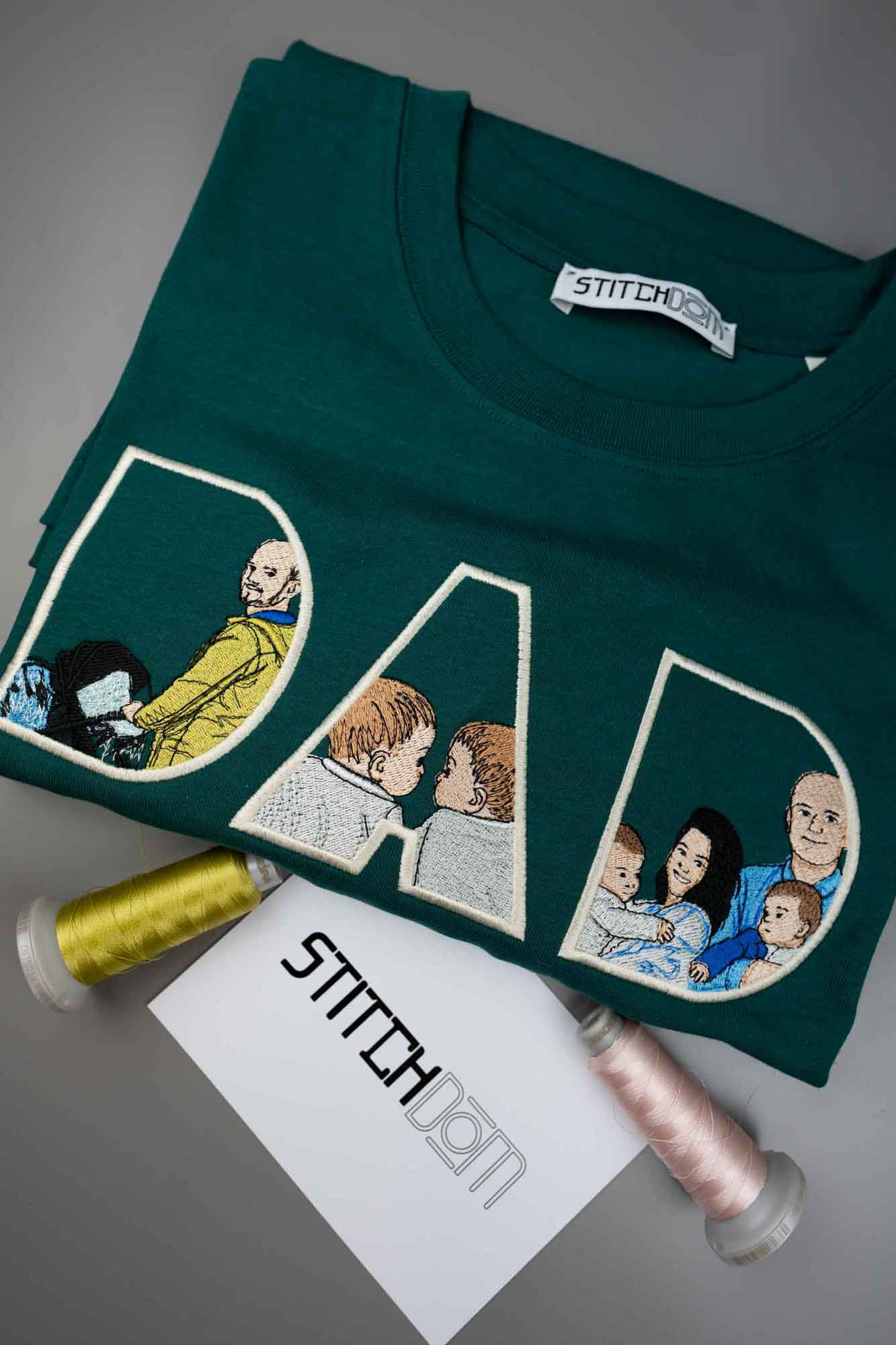 Tricou verde închis petrol împăturit, cu textul „DAD” brodat în litere mari conturate, fiecare literă conținând portrete brodate ale familiei, pe o suprafață gri, alături de două conuri de ață și un card branduit Stitchdom.