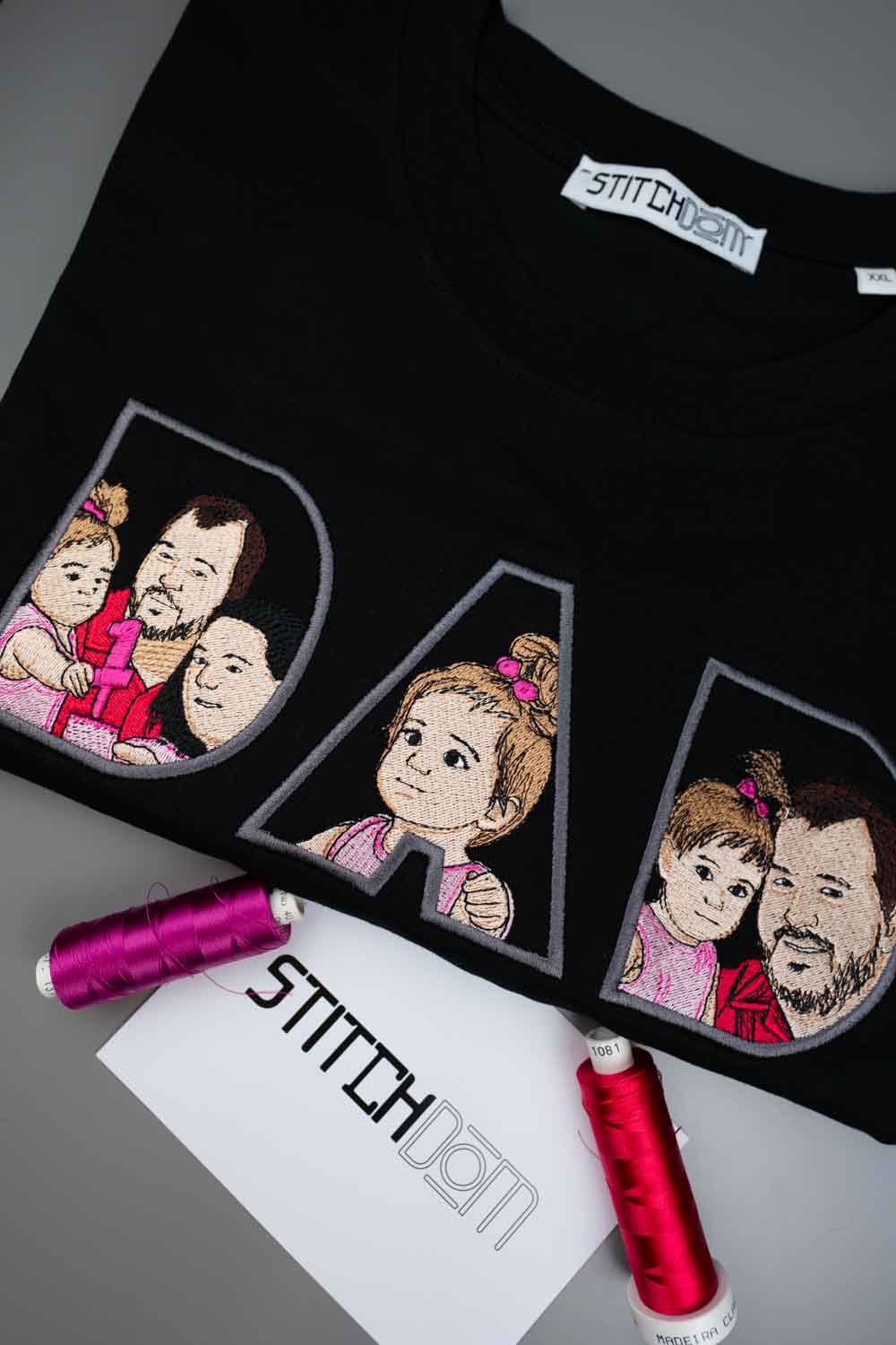 Tricou negru din bumbac organic, împăturit, cu textul „DAD” realizat din trei litere mari conturate, fiecare literă având în interior o poză de familie brodată, pe o suprafață gri, cu două conuri de ață și un card de brand „Stitchdom” în față.