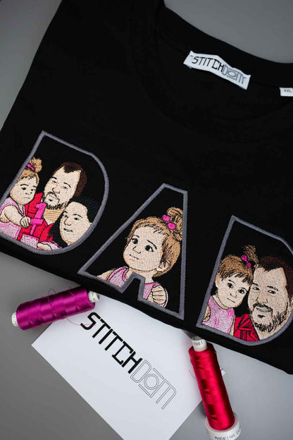 Tricou negru din bumbac organic, împăturit, cu textul „DAD” realizat din trei litere mari conturate, fiecare literă având în interior o poză de familie brodată, pe o suprafață gri, cu două conuri de ață și un card de brand „Stitchdom” în față.