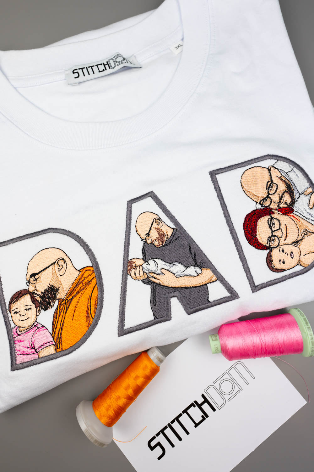 Tricou alb din bumbac organic, cu textul „DAD” realizat din litere mari brodate, fiecare literă având în interior o poză brodată personalizată în stil desen cu un tată și copilul său, pe o suprafață gri, alături de conuri de ață colorate și un card de brand „Stitchdom”.