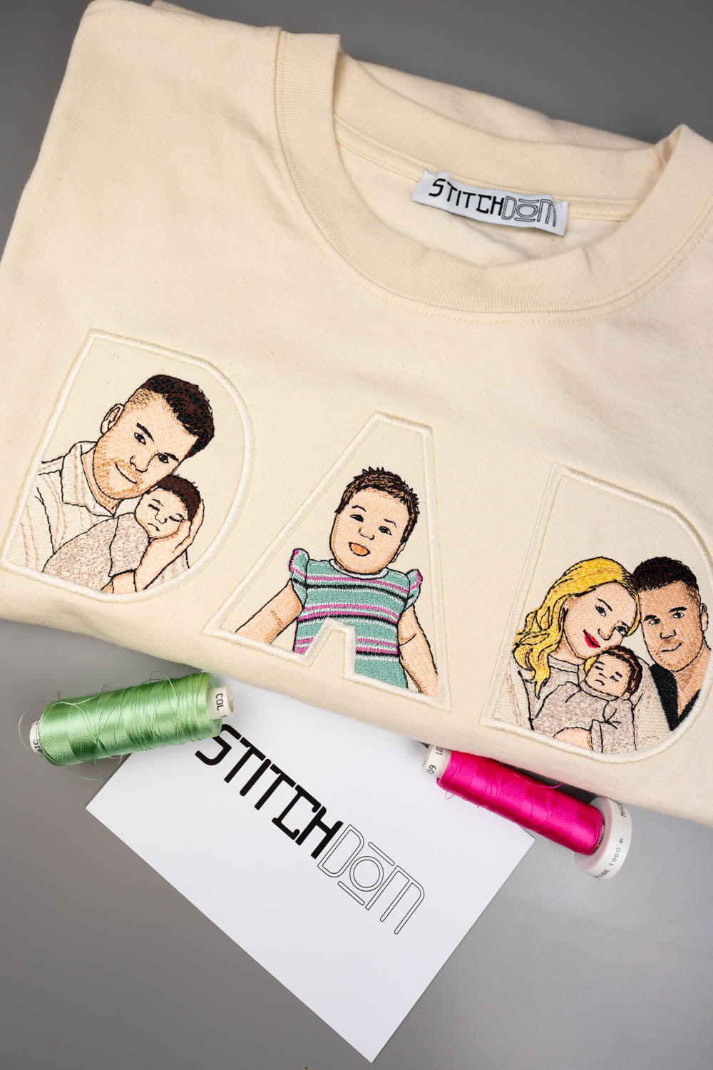 Tricou crem din bumbac organic, împăturit, cu textul „DAD” realizat din litere brodate, fiecare literă având în interior poze de familie brodate personalizate, alături de două conuri de ață și un card de brand, pe un fundal gri.