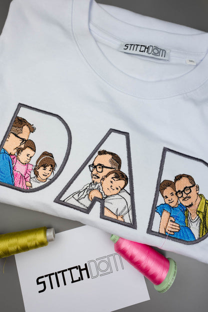 Tricou alb din bumbac organic, așezat pe o suprafață gri, cu textul „DAD” realizat din litere mari conturate și umplute cu poze de familie brodate în stil desen, alături de două conuri de ață și un card de brand „StitchDOM”.