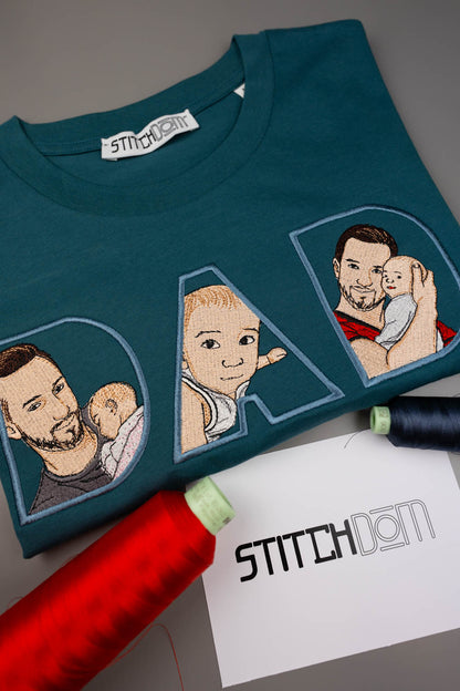 Tricou albastru închis din bumbac organic, împăturit, cu literele „DAD” brodate personalizat, având în interior poze brodate cu un tată și copilul său, alături de conuri de ață și un card de brand „Stitchdom”, pe un fundal gri.