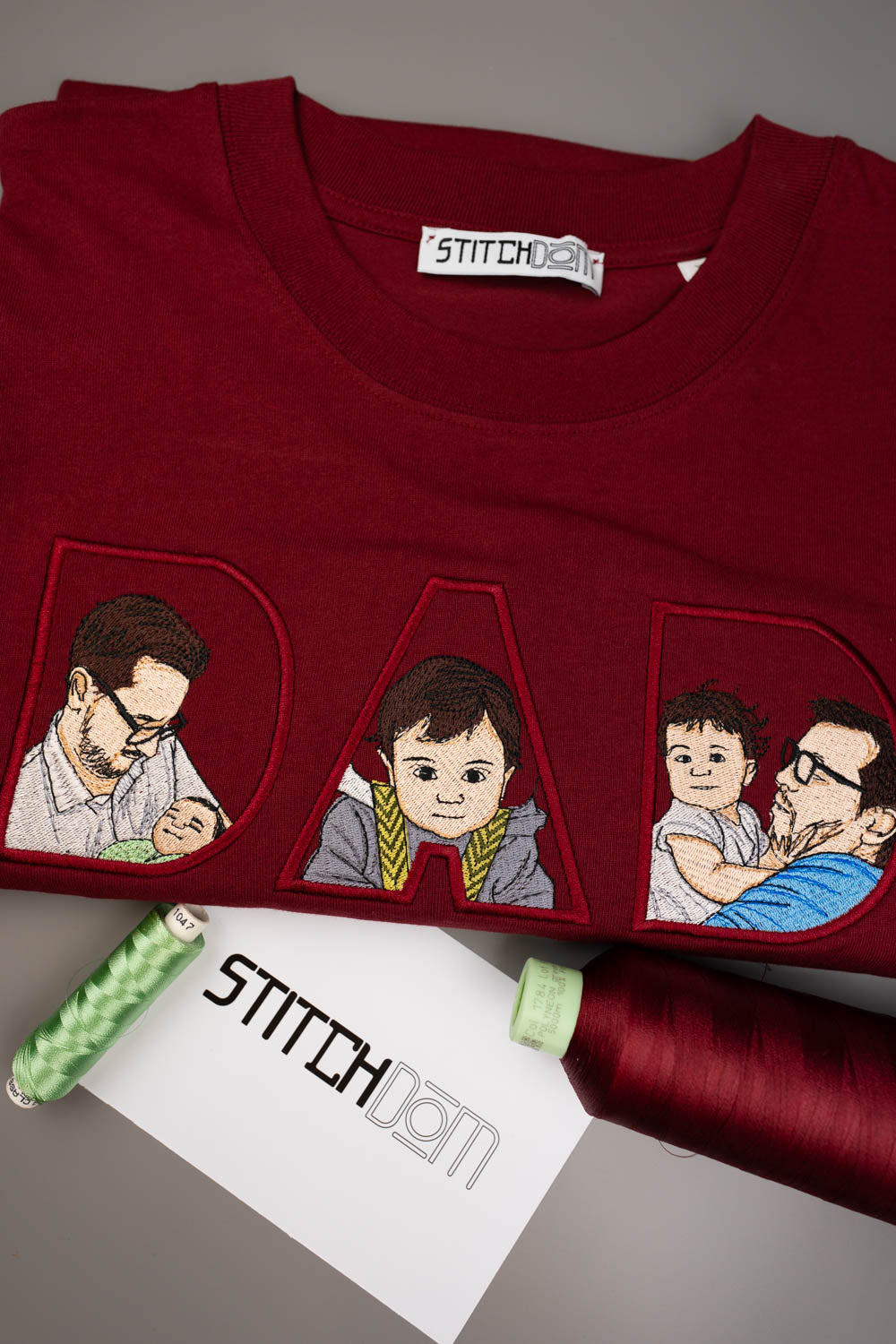 Tricou roșu închis din bumbac organic, împăturit, cu literele „DAD” brodate personalizat, având în interior poze brodate cu un tată și copilul său, alături de conuri de ață roșie și verde și o etichetă de brand „Stitchdom”, pe un fundal gri.