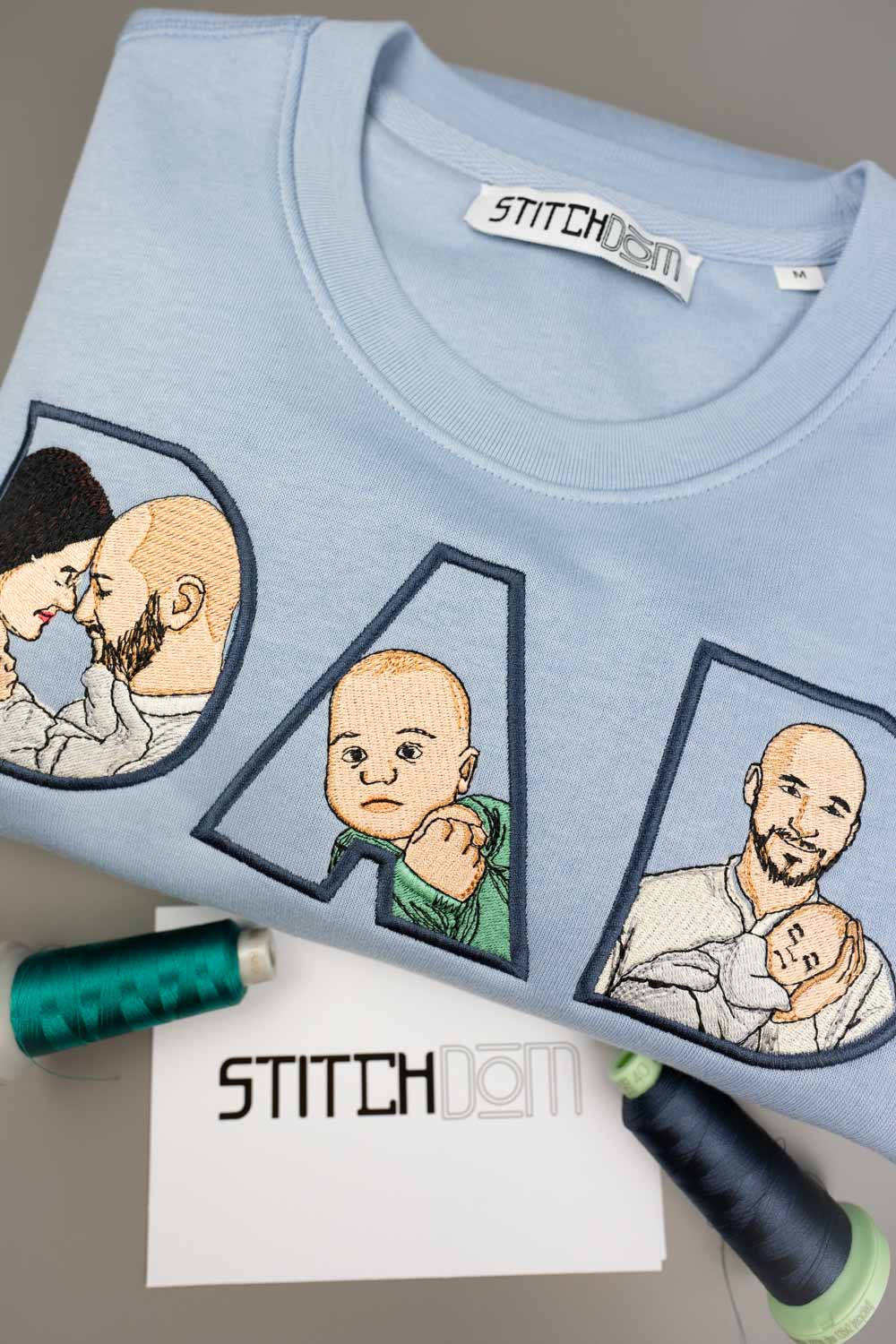 Tricou bleu deschis din bumbac organic, împăturit, de la Stitchdom, cu literele „D”, „A” și „M” brodate personalizat, având în interior poze brodate cu un tată și copilul său, pe o suprafață gri, alături de conuri de ață și o etichetă albă.