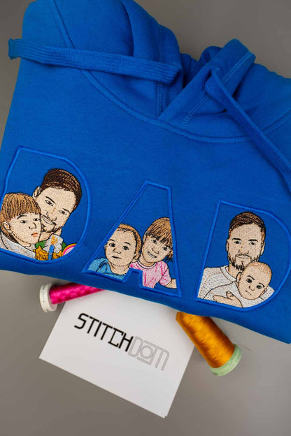 Hanorac bleumarin închis din bumbac organic, împăturit, cu poze de familie brodate în interiorul literelor „DAD”, pe o suprafață gri, alături de un card alb cu textul „STITCHDOM” și două conuri de ață.
