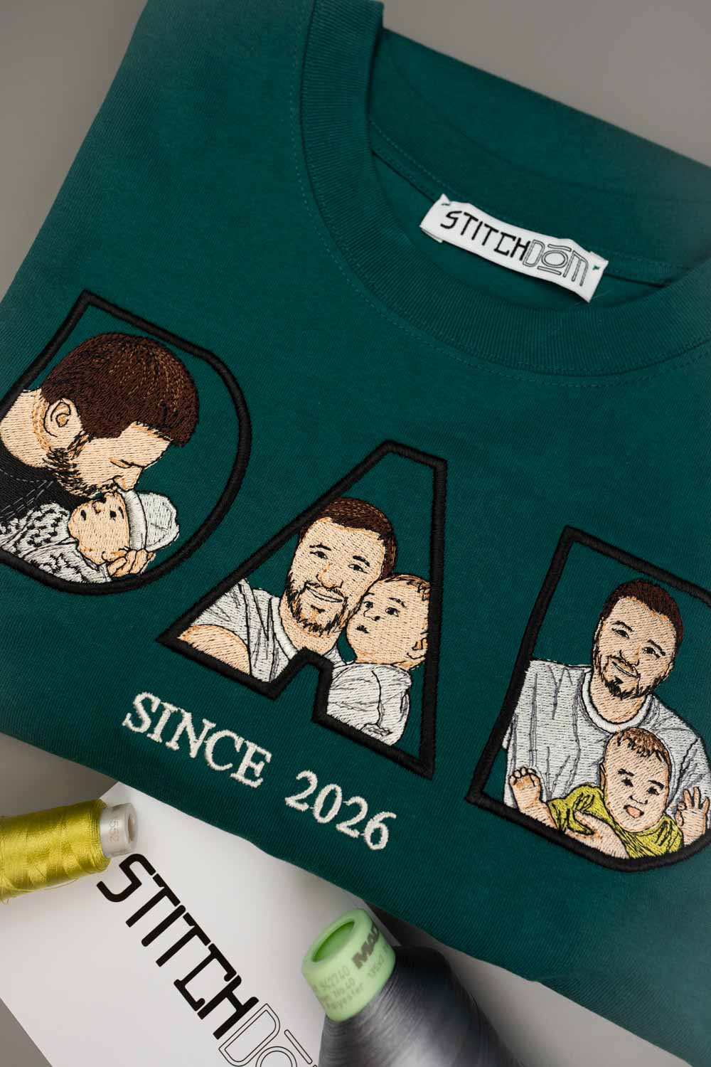 Tricou verde petrol închis din bumbac organic, împăturit, cu textul „DAD” brodat personalizat, cu poze de familie brodate în interiorul literelor, textul „SINCE 2026” și eticheta de brand „Stitchdom”.
