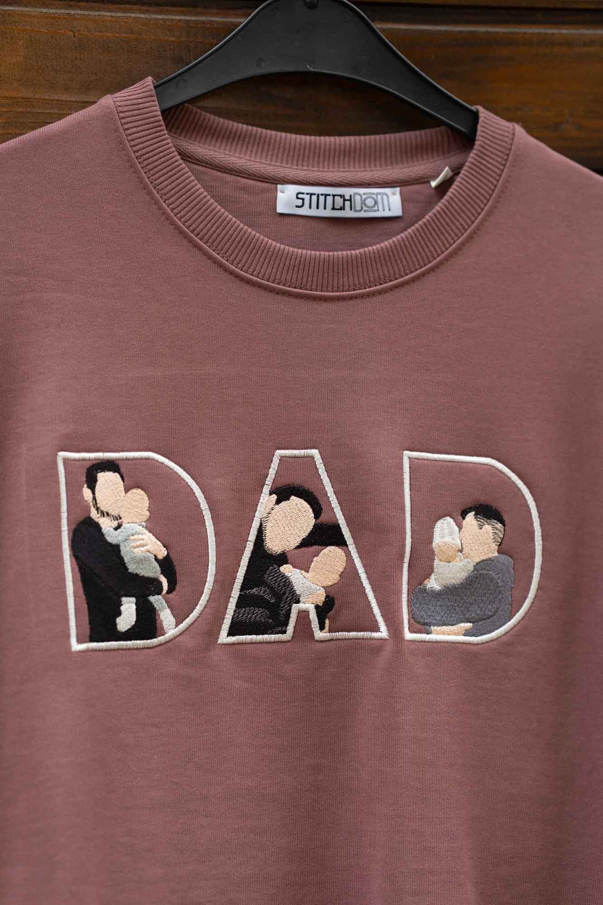 Hanorac maro cu guler rotund, pe umeraș negru, pe fundal din lemn, cu textul „DAD” brodat, fiecare literă conținând fotografii brodate ale familiei.