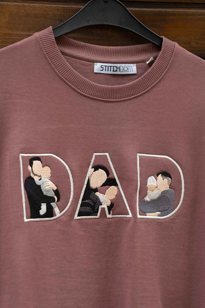Hanorac maro cu guler rotund, pe umeraș negru, pe fundal din lemn, cu textul „DAD” brodat, fiecare literă conținând fotografii brodate ale familiei.