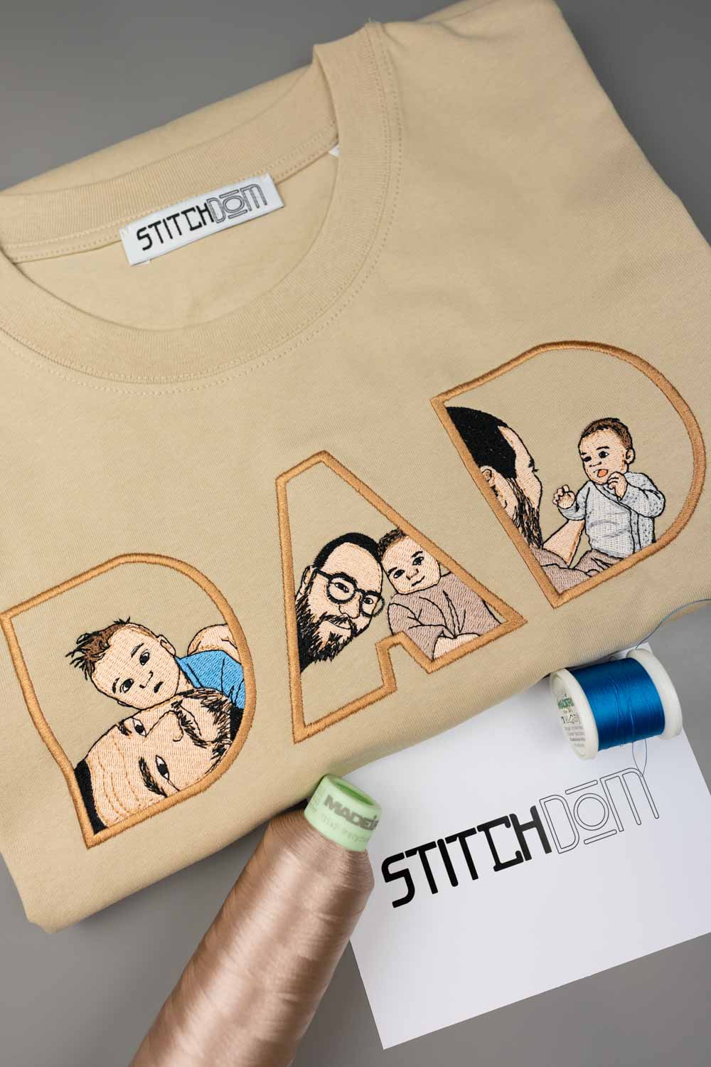 Tricou bej din bumbac organic, cu textul „DAD” realizat din litere mari brodate, fiecare literă având în interior o poză brodată cu un tată și copilul său, alături de conuri de ață și un card de brand „Stitchdom”, pe un fundal gri.