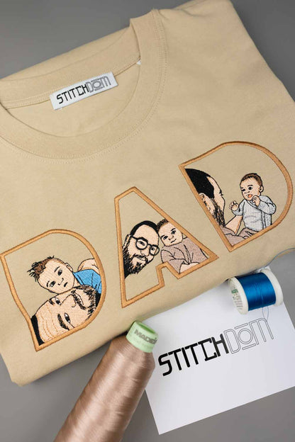 Tricou bej din bumbac organic, cu textul „DAD” realizat din litere mari brodate, fiecare literă având în interior o poză brodată cu un tată și copilul său, alături de conuri de ață și un card de brand „Stitchdom”, pe un fundal gri.