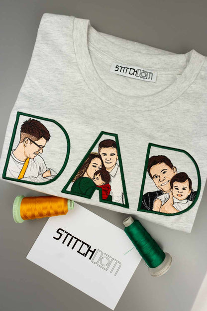 Tricou gri deschis din bumbac organic, împăturit, cu textul „DAD” brodat personalizat, cu poze de familie brodate în interiorul literelor, pe o suprafață gri, alături de conuri de ață și un card „Stitchdom”.