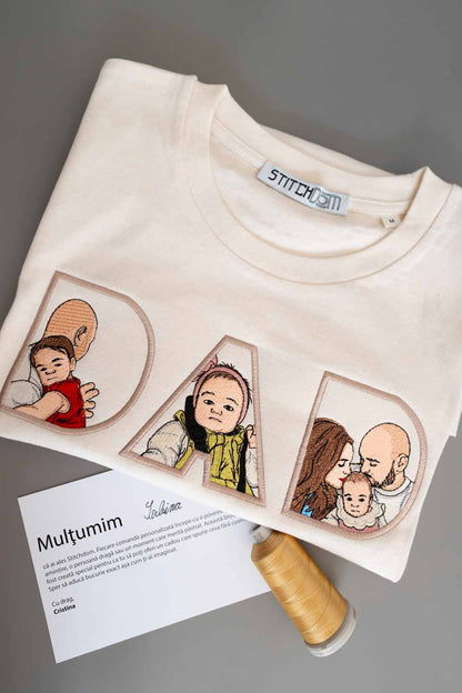 Tricou crem din bumbac organic, împăturit, cu o poză brodată personalizată cu o familie pe partea din față, pe un fundal gri, alături de un con de ață și un card imprimat cu textul „Mulțumim”.