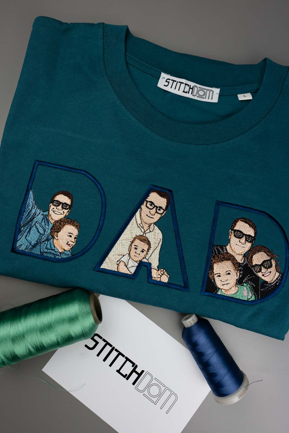 Tricou verde petrol din bumbac organic, împăturit, cu textul „DAD” realizat din litere mari brodate, fiecare literă având în interior portrete de familie brodate personalizat, alături de conuri de ață verde, albă și bleu și un card alb cu logo-ul „STITCHDOM”, pe un fundal gri.