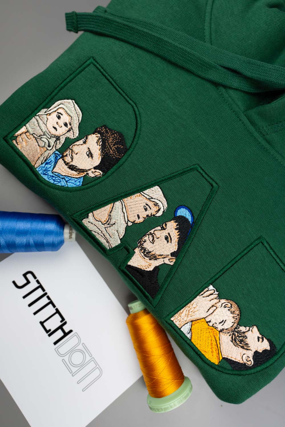 Hanorac verde din bumbac organic, împăturit, cu textul „DAD” realizat din litere mari brodate, fiecare literă având în interior portrete de familie brodate personalizat, alături de conuri de ață albastra si portocalie și un card alb cu logo-ul „STITCHNOM”, pe un fundal gri.