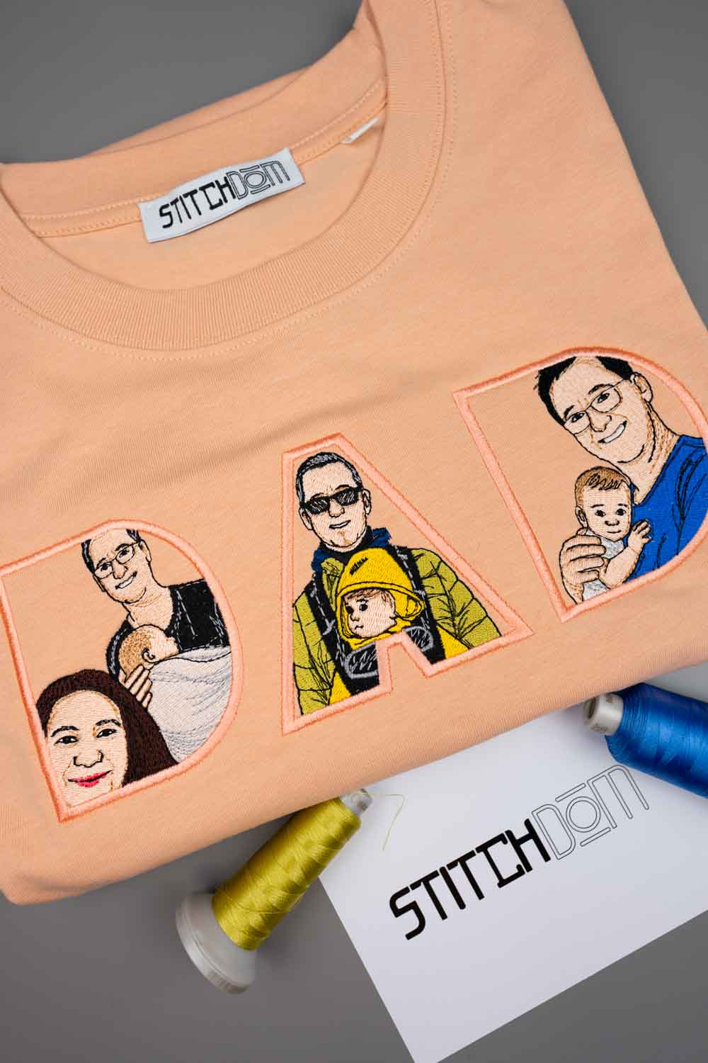 Tricou piersică din bumbac organic, cu guler rotund, cu textul „DAD” realizat din litere brodate personalizat, fiecare literă având în interior un portret de familie în stil desen, pe un fundal gri, alături de conuri de ață și o etichetă de brand „StitchDOM”.