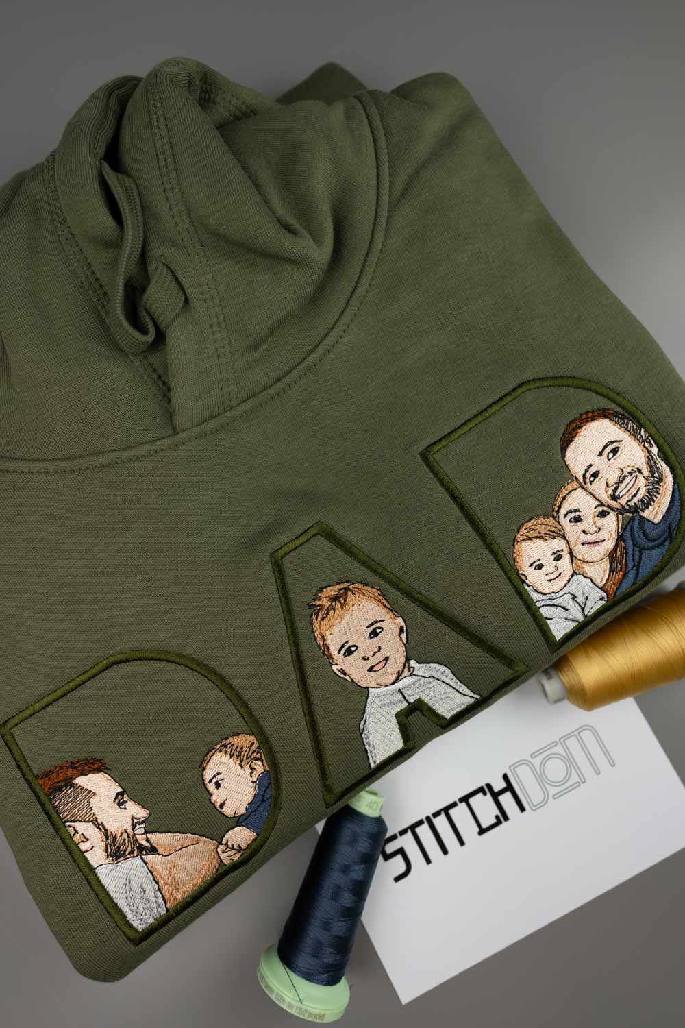 Hanorac verde măsliniu din bumbac organic, așezat flat lay pe un fundal gri, cu portrete de familie brodate în interiorul literelor „DAD”, alături de conuri de ață albastră și aurie și un card cu textul „STITCHDOM”.