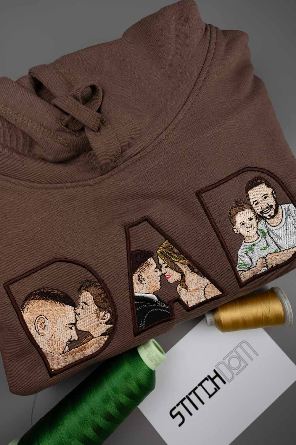 Hanorac maro din bumbac organic, împăturit, cu literele „DAD” brodate, fiecare literă având un portret brodat în stil patch, pe un fundal gri, alături de conuri de ață și un card cu textul „STITCHDOM”.