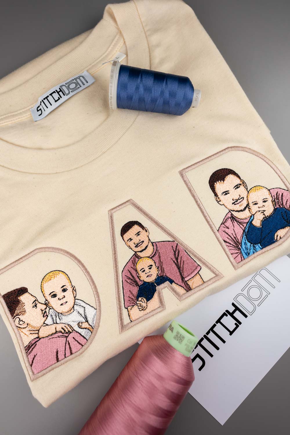 Tricou crem din bumbac organic, împăturit, cu portrete de familie brodate în interiorul literelor „DAD”, alături de conuri de ață albastră și roz și o etichetă de brand „Stitchdom”.