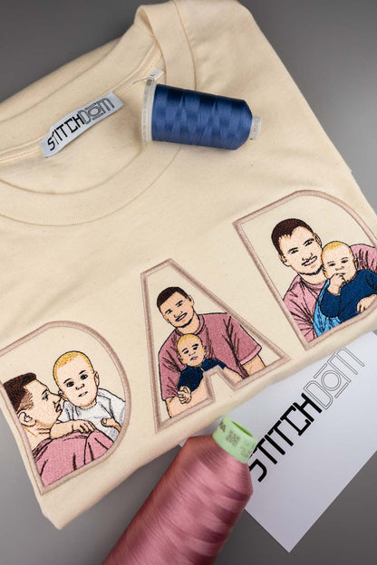 Tricou crem din bumbac organic, împăturit, cu portrete de familie brodate în interiorul literelor „DAD”, alături de conuri de ață albastră și roz și o etichetă de brand „Stitchdom”.