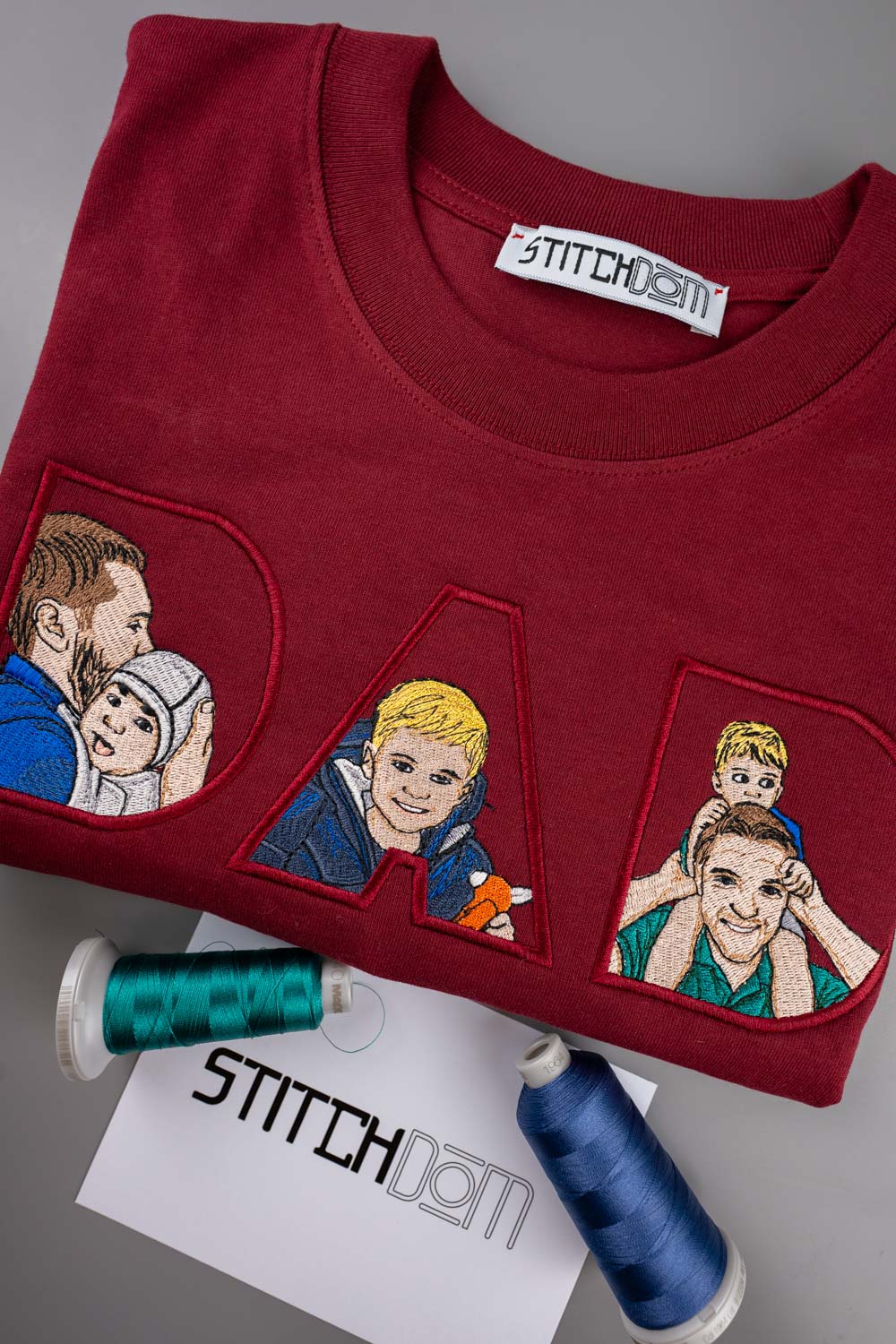 Tricou bordo din bumbac organic, cu guler rotund, împăturit, cu literele mari „D”, „A” și „D” brodate în stil cartoon pe piept, pe un fundal gri, alături de două conuri de ață și o etichetă de brand „Stitchdom”.
