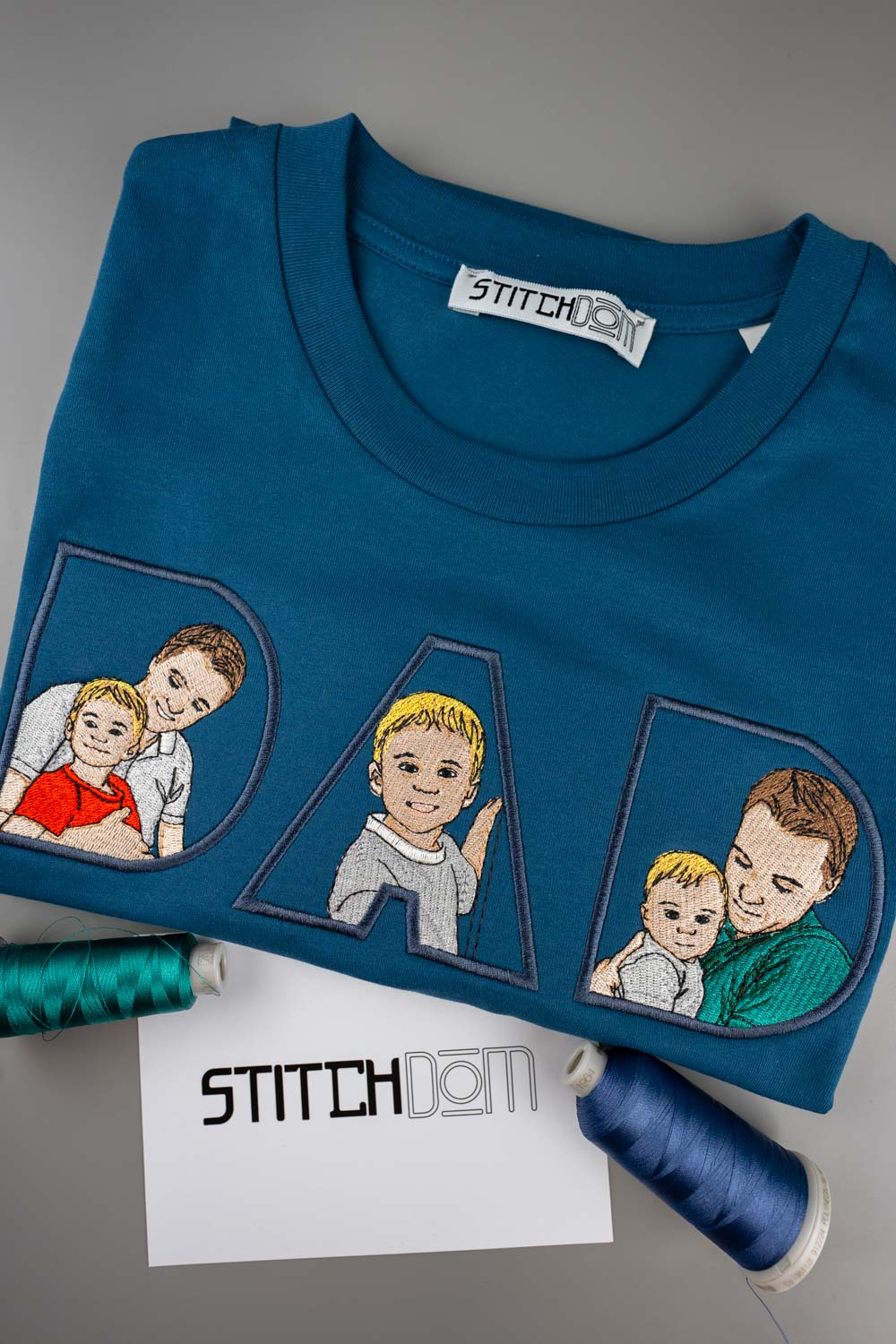 Tricou albastru din bumbac organic, împăturit, cu literele „DAD” brodate, având în interior portrete ilustrate brodate ale familiei, pe un fundal gri, alături de conuri de ață și un card cu brandul „Stitchdom”.
