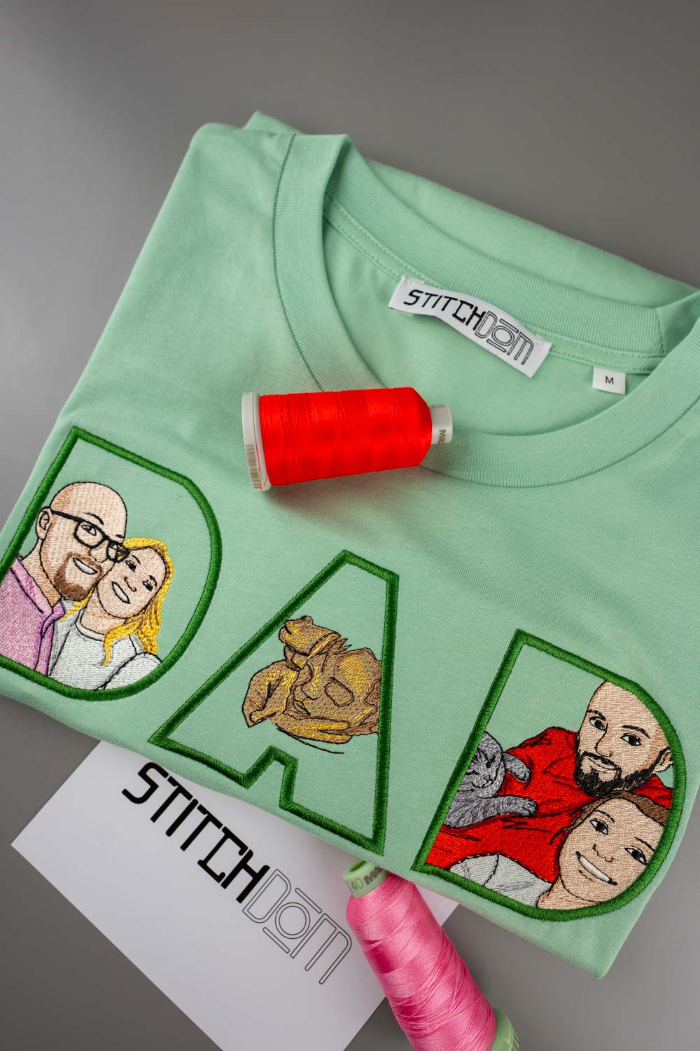 Tricou verde mentă din bumbac organic, cu literele „DAD” brodate, având în interior portrete brodate ale unui cuplu și ale unui câine, alături de conuri de ață roșie și roz, pe un fundal gri.