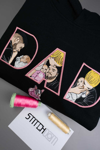 Hanorac negru din bumbac organic, împăturit, cu literele mari „DAD” brodate în roz, având portrete ilustrate brodate cu tați și copii în interiorul literelor, pe o suprafață gri, alături de două conuri de ață și un card cu textul „STITCHDOM”.