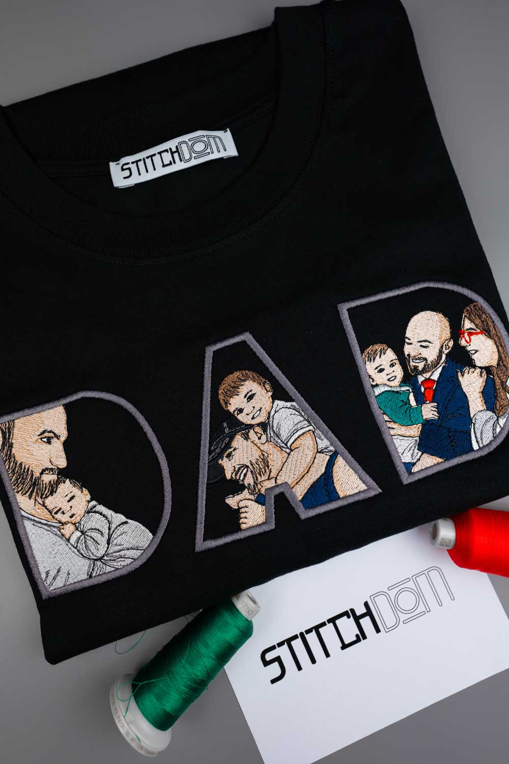 Tricou negru din bumbac organic, împăturit, cu literele „DAD” brodate, fiecare literă având portrete de familie brodate în stil patch, pe un fundal gri, alături de conuri de ață și un card cu brandul „Stitchdom”.