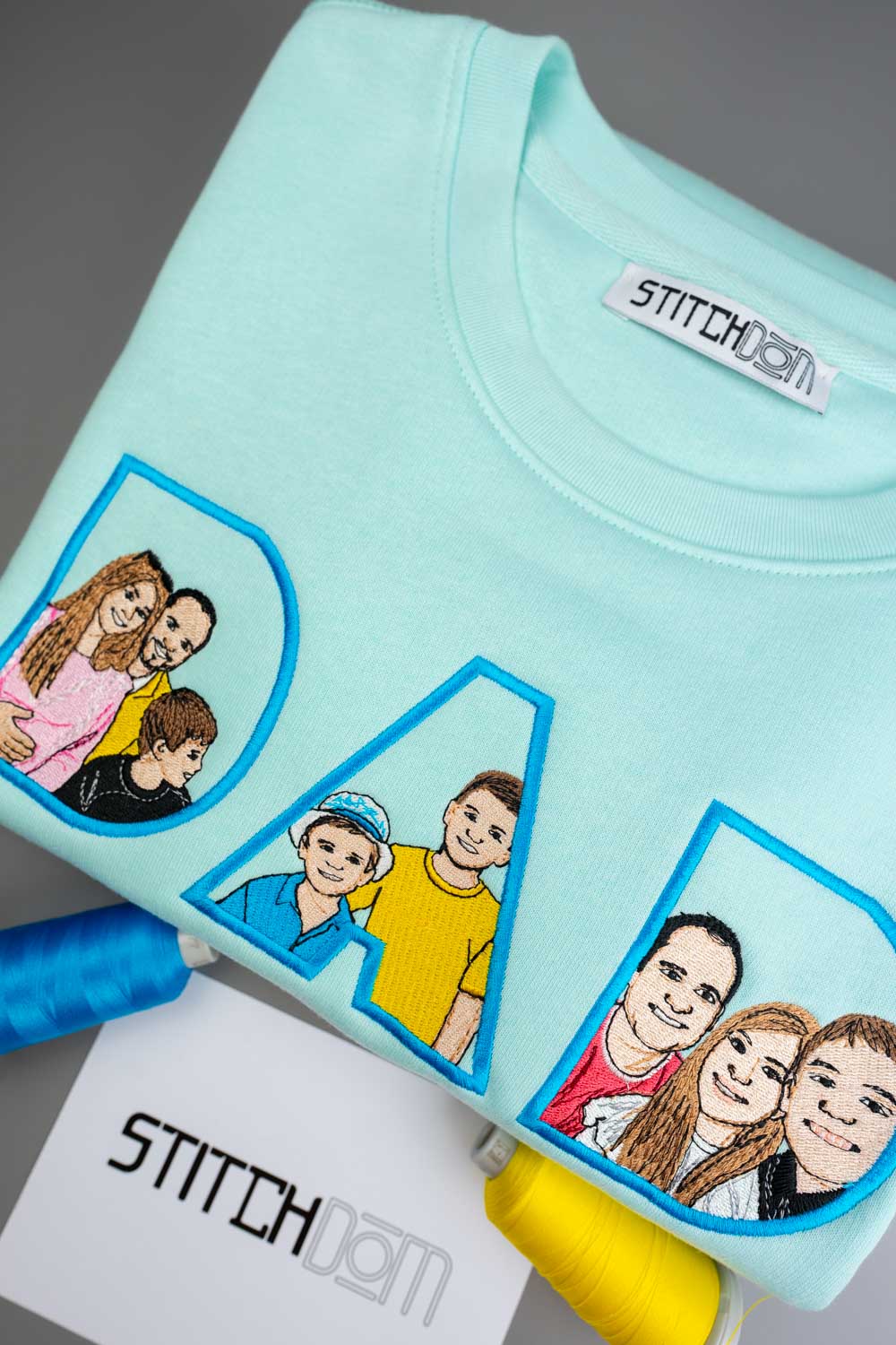 Bluza verde mentă din bumbac organic, împăturit, cu portrete de familie brodate în interiorul literelor „DAD” pe partea din față, cu brandul „STITCHDOM” vizibil pe eticheta interioară de la gât și pe un card în prim-plan.
