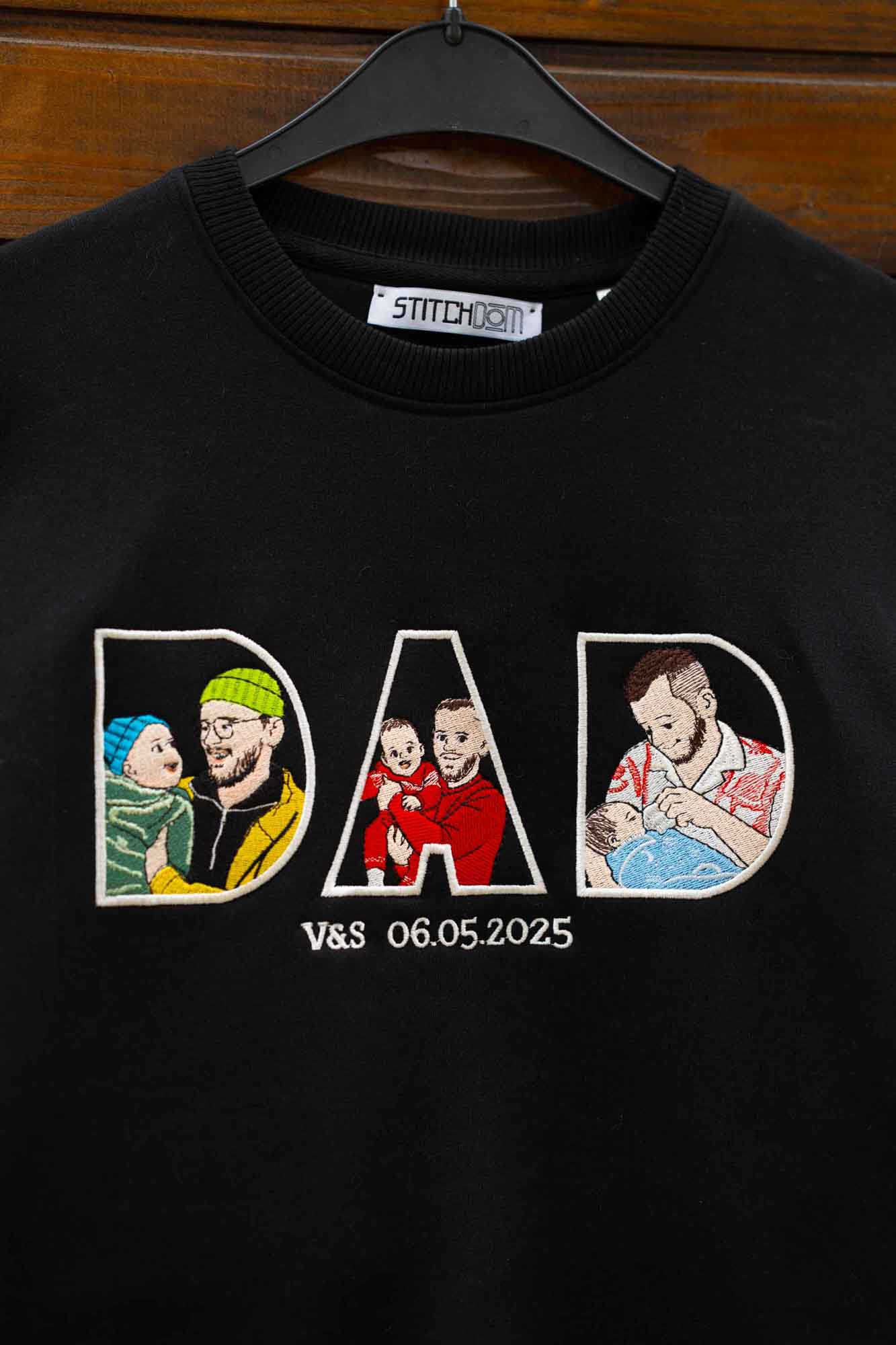 Hanorac negru cu guler rotund, pe umeraș negru, pe fundal din lemn, cu textul „DAD” brodat, fiecare literă conținând fotografii brodate ale familiei, și textul „V&S 06”.