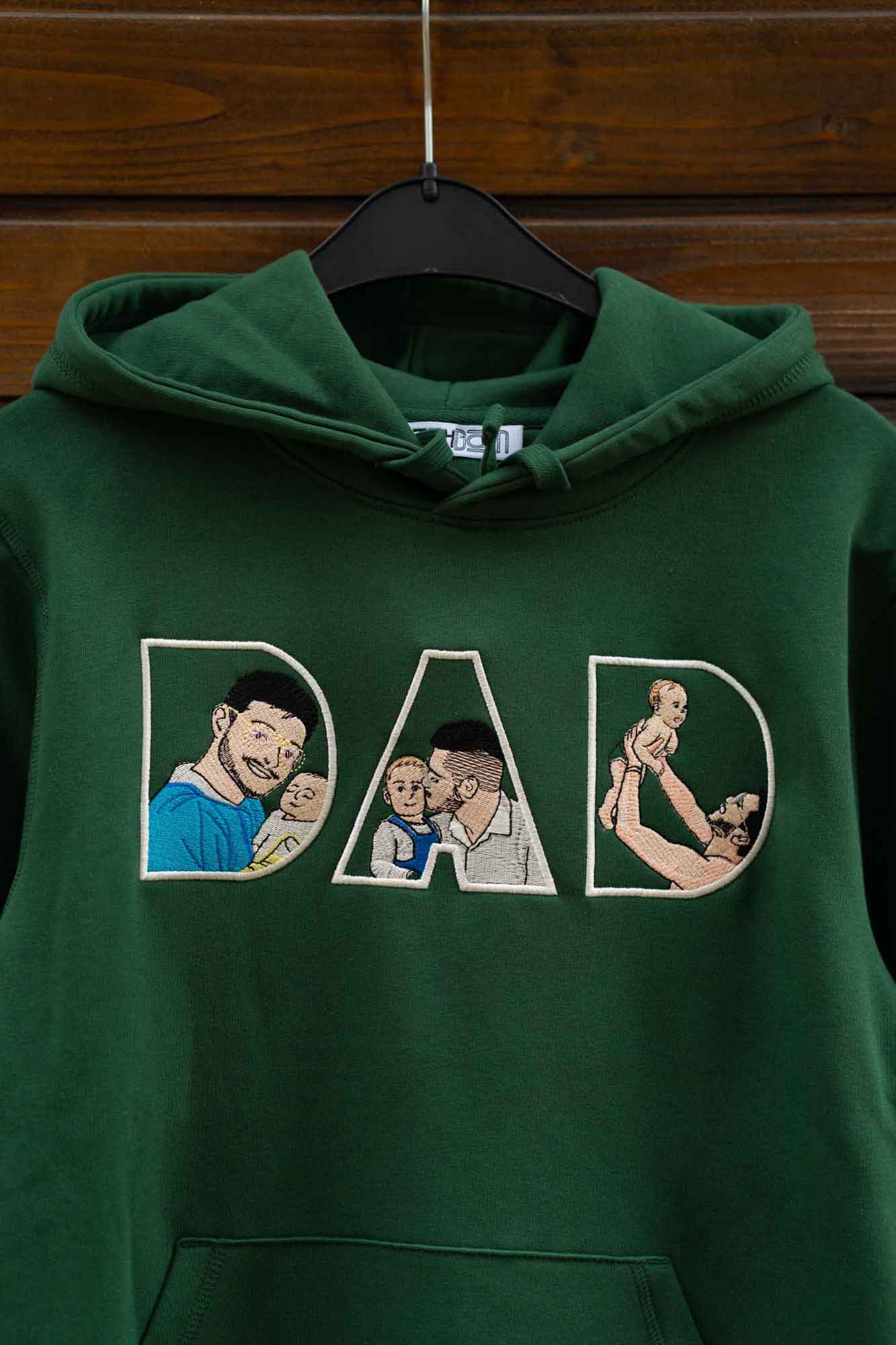 Hanorac verde cu glugă, pe umeraș, cu textul „DAD” brodat, fiecare literă conținând elemente brodate, pe fundal din lemn.
