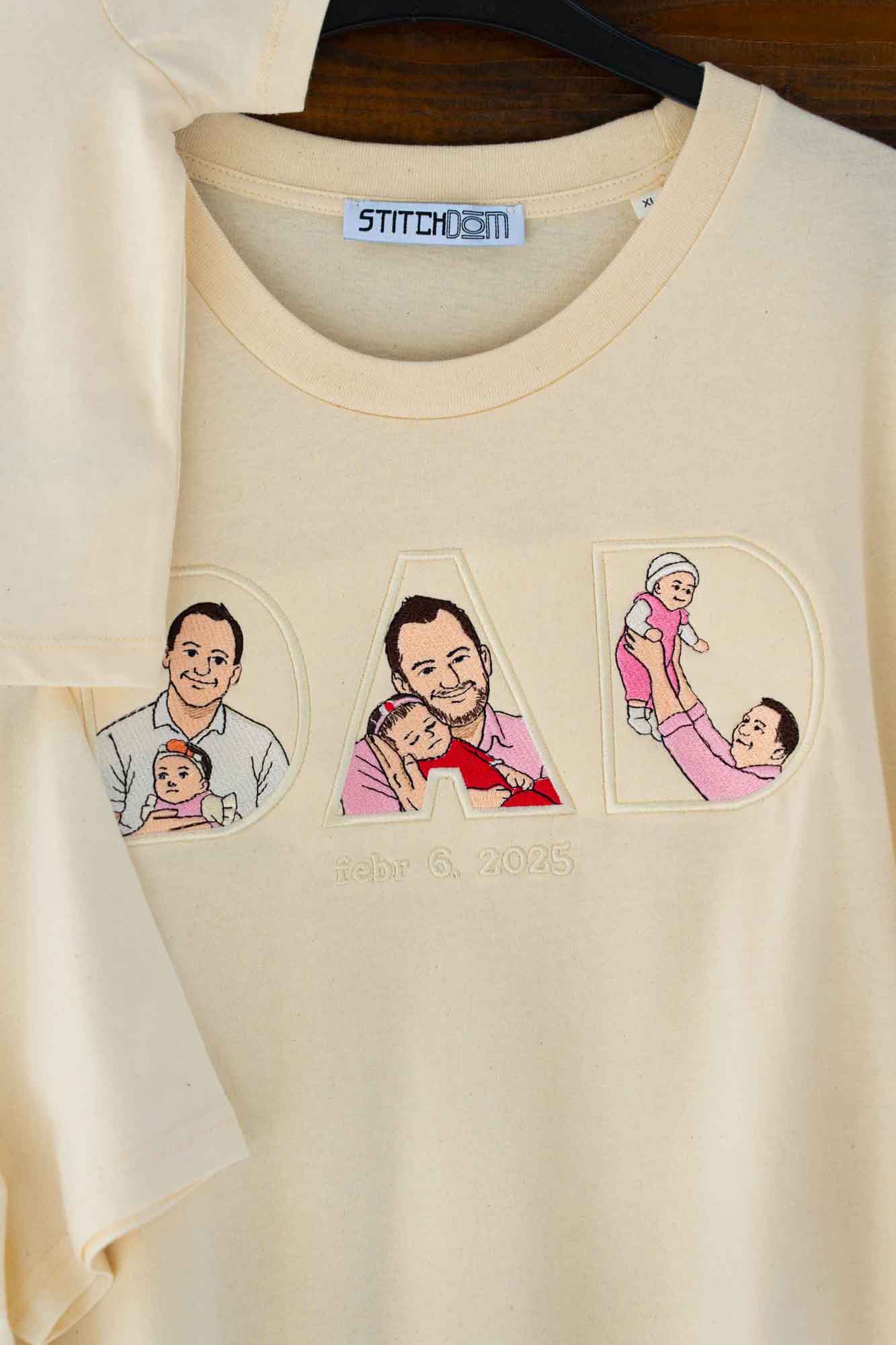 Tricou bej cu textul „DAD” brodat, fiecare literă conținând imagini brodate reprezentând familia, pe o suprafață din lemn.