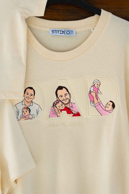 Tricou bej cu textul „DAD” brodat, fiecare literă conținând imagini brodate reprezentând familia, pe o suprafață din lemn.