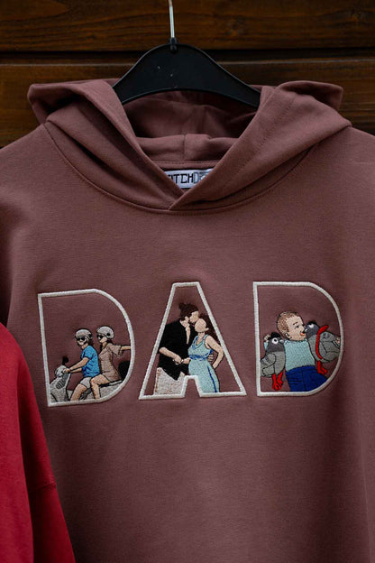 Hanorac maro cu glugă, pe umeraș, cu textul „DAD” brodat, fiecare literă conținând imagini brodate reprezentând un tată și un copil.