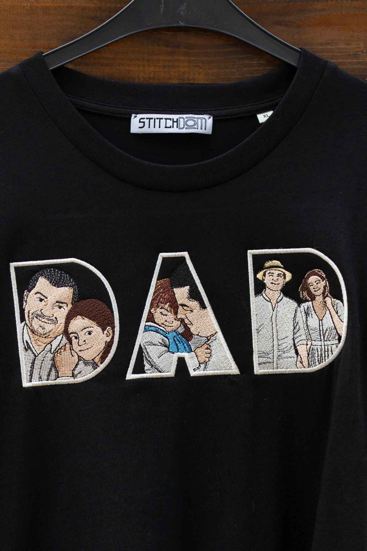 Tricou negru cu guler rotund, pe umeraș negru, pe fundal din lemn închis, cu un model brodat mare reprezentând textul „DAD” pe piept, fiecare literă conținând câte un panou cu portrete ilustrate ale unui cuplu.