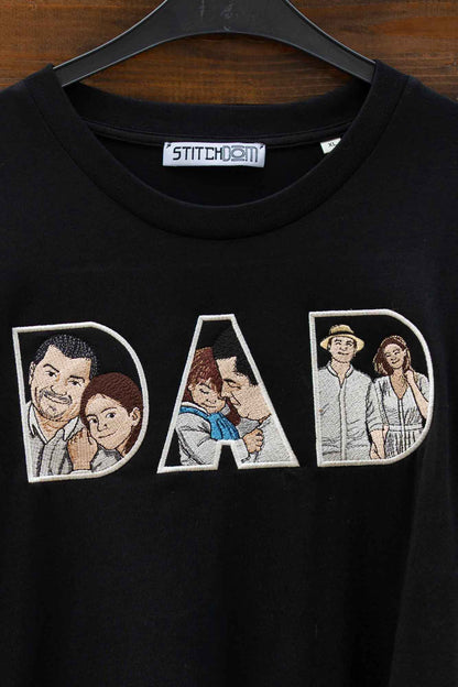 Tricou negru cu guler rotund, pe umeraș negru, pe fundal din lemn închis, cu un model brodat mare reprezentând textul „DAD” pe piept, fiecare literă conținând câte un panou cu portrete ilustrate ale unui cuplu.