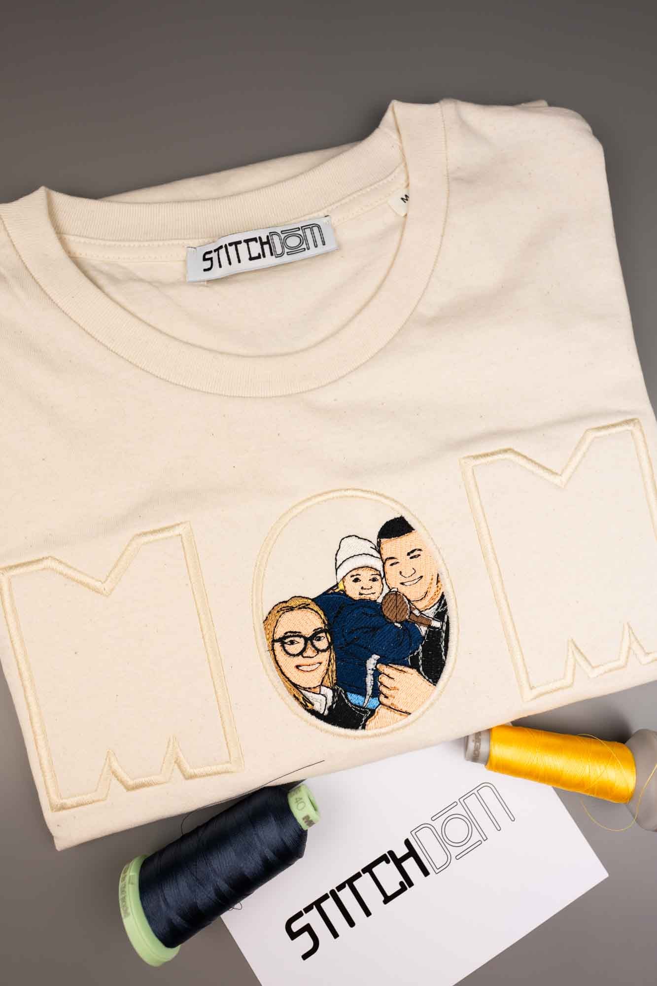 Un tricou crem, din bumbac organic, împăturit, cu un design brodat personalizat ce reprezintă cuvântul „MOM” în litere mari conturate, având în centru un portret oval al unei familii. Tricoul este prezentat pe un fundal gri, alături de conuri de ață și un cartonaș Stitchdom.