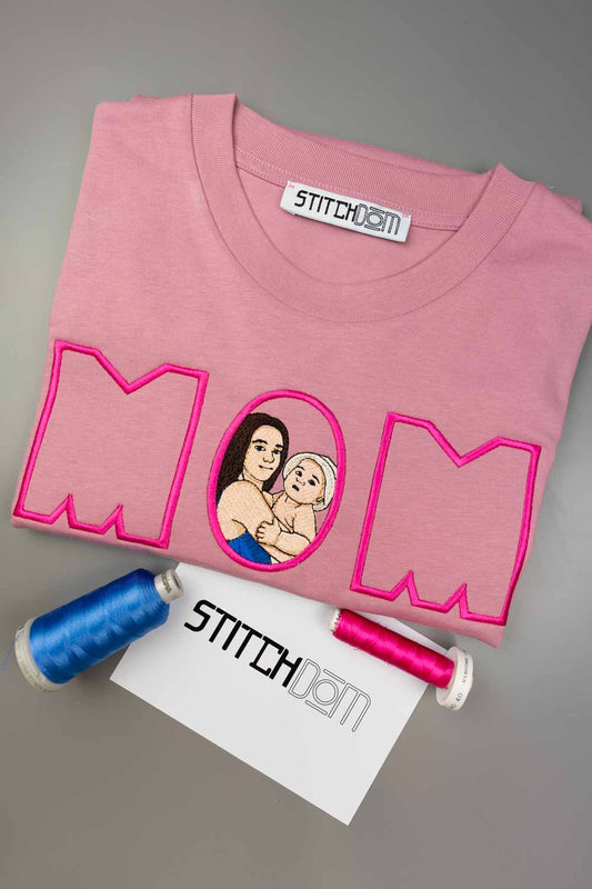 Tricou roz din bumbac organic, împăturit, cu textul „MOM” brodat cu roz aprins și o poză centrală în stil desen cu o mamă și copilul său, alături de două conuri de ață, unul bleu și unul roz, și un card alb cu logo-ul „Stitchdom”, pe un fundal gri.