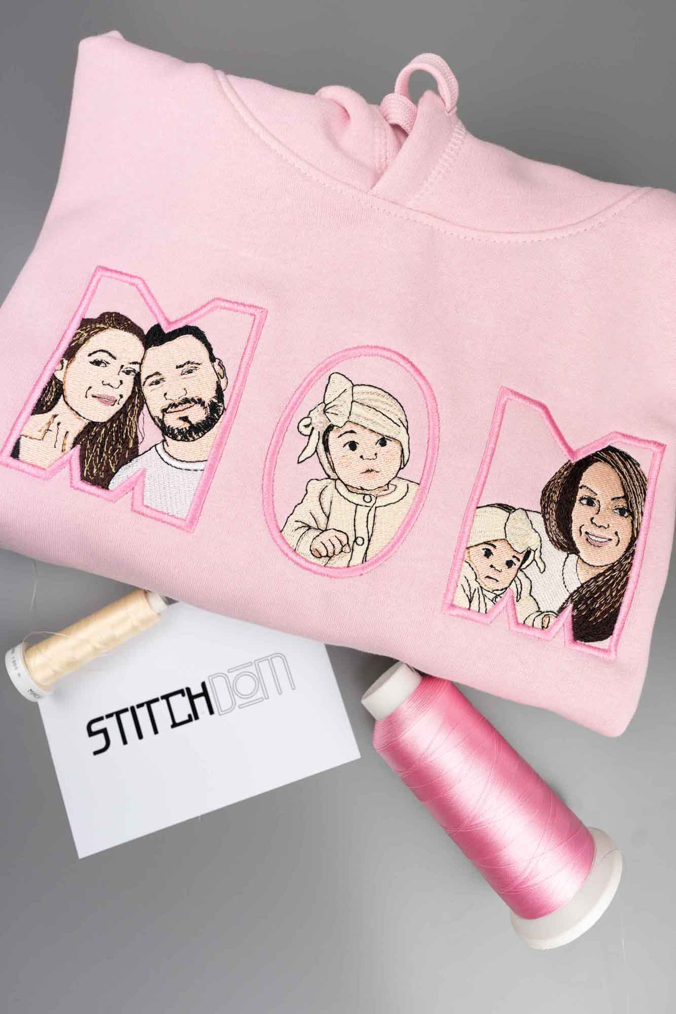 Hanorac roz cu textul „MOM” brodat, fiecare literă conținând portrete brodate reprezentând familia, pe un fundal gri, cu un card branduit Stitchdom și conuri de ață roz vizibile.